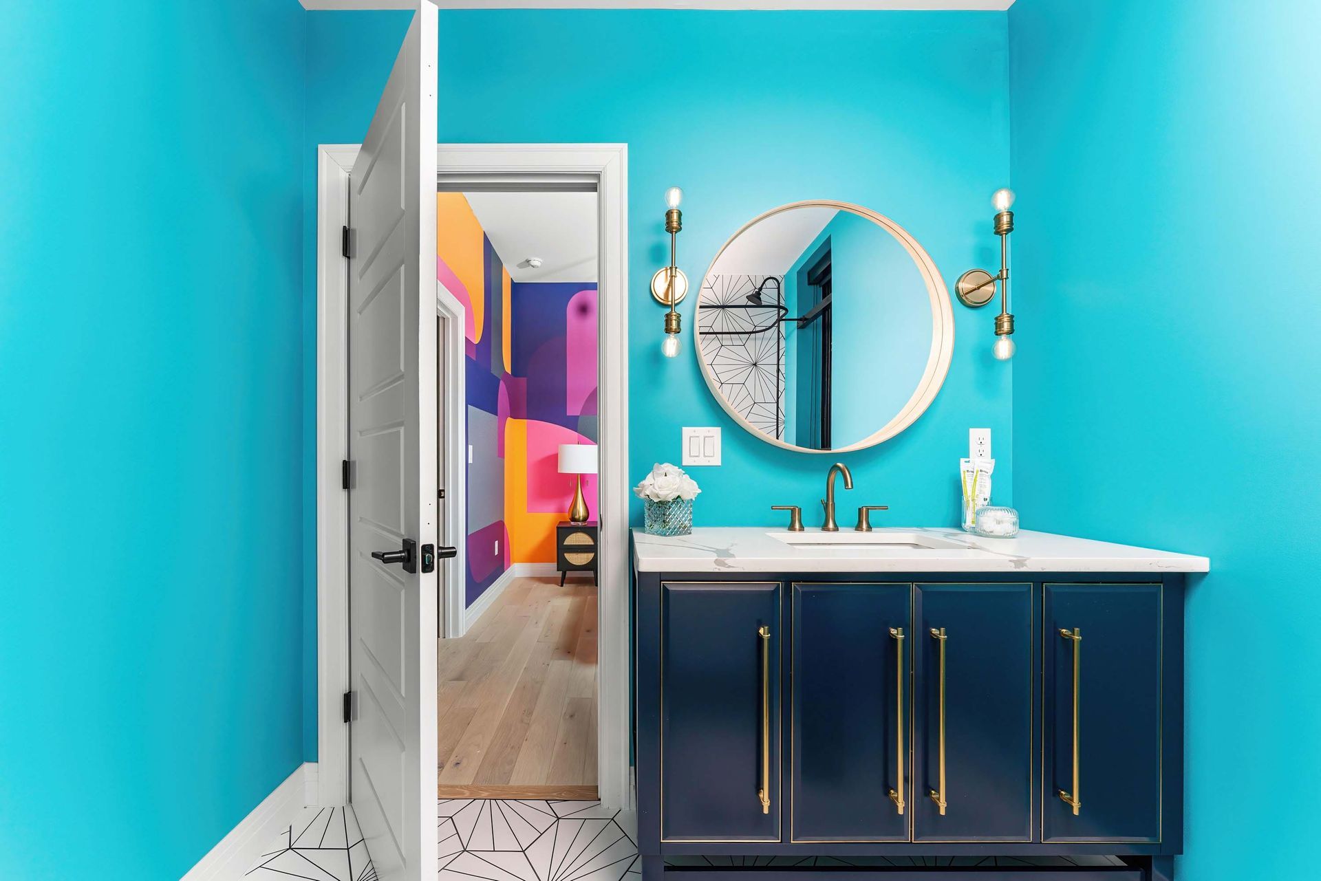Bright blue bathroom with navy vanity, round mirror, and open door to a colorful hallway