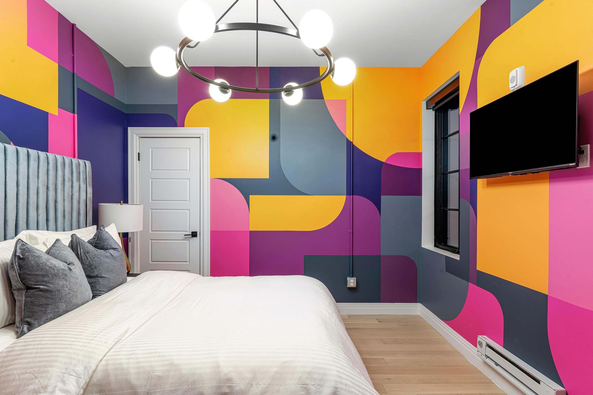 Colorful bedroom with bold geometric wall art, white bed, ceiling light, and wall-mounted TV