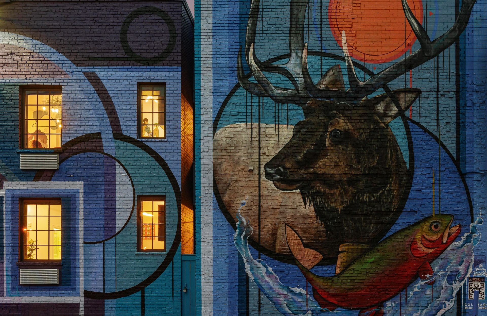 Mural of a deer and red fish on a brick building at dusk, with glowing windows and blue-orange abstract swirls