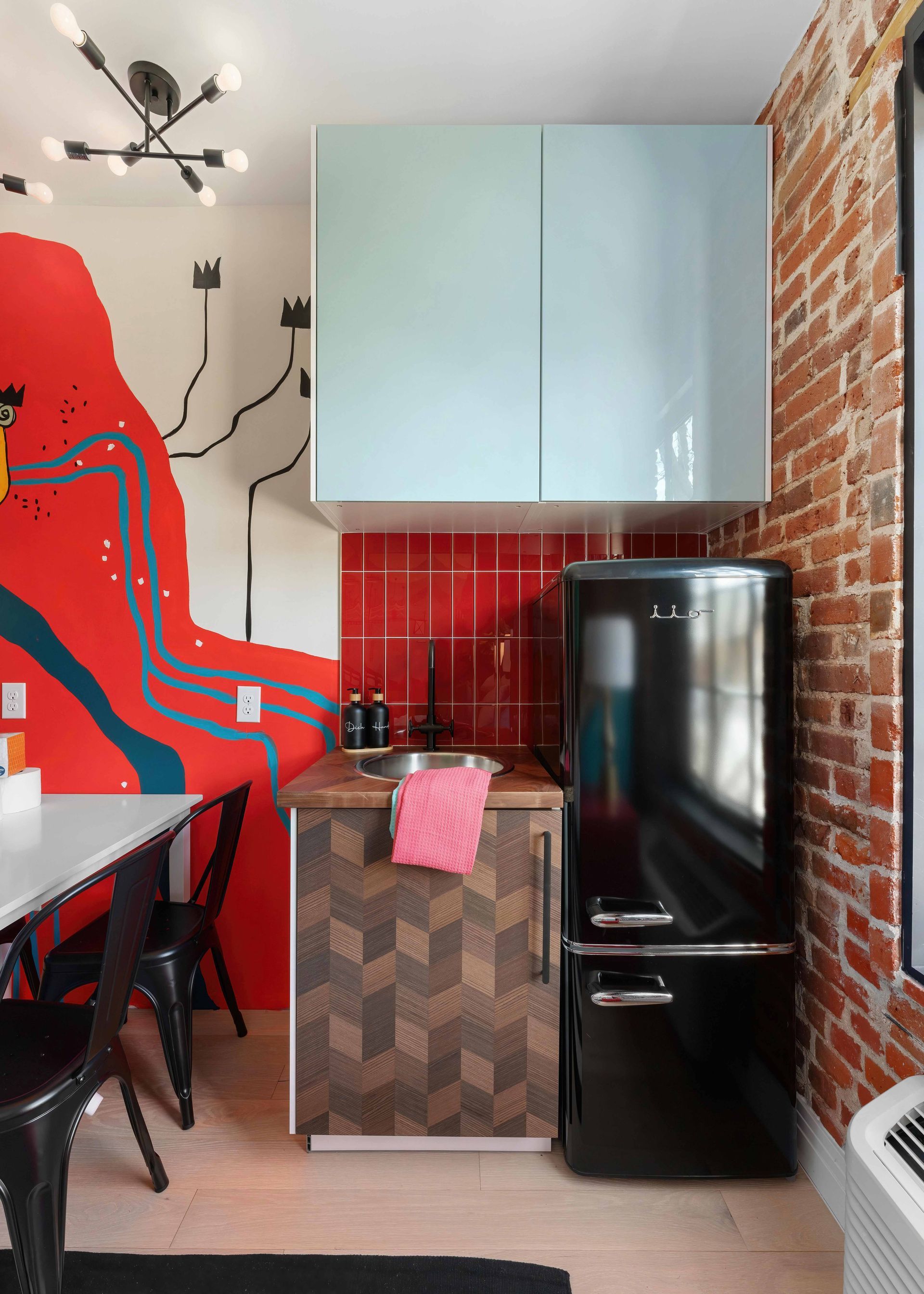 Small kitchen with red tile backsplash, pale blue cabinets, black fridge, and brick wall beside a dining area.