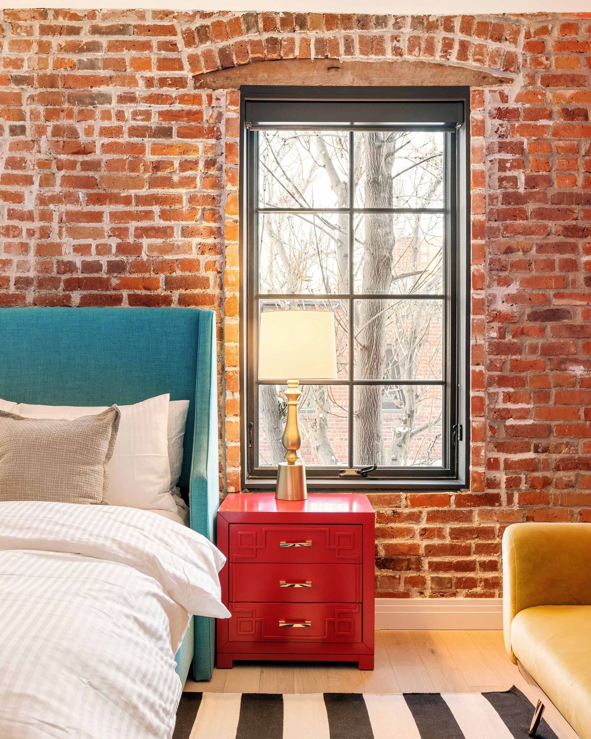 Brick-walled bedroom with teal bed, red nightstand, and mustard chair by a window