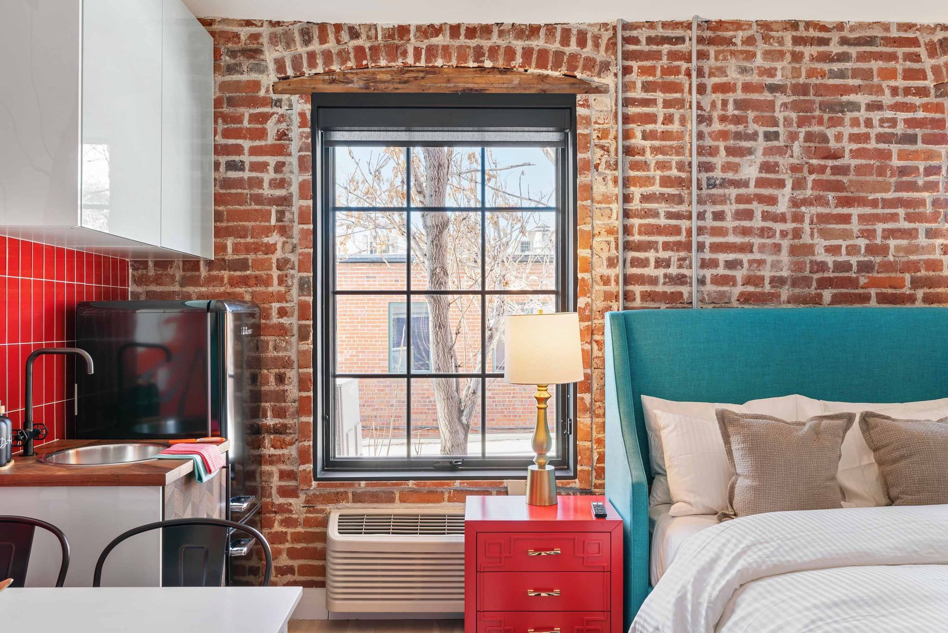 Loft bedroom with exposed brick wall, teal bed, red nightstand, and a black-framed window