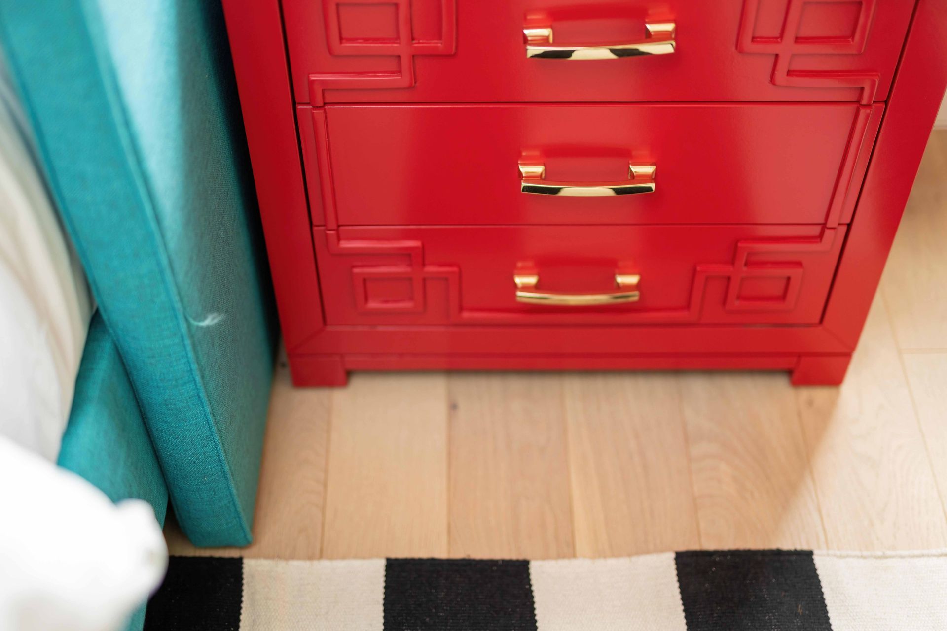 Red dresser beside teal chair, with striped rug on light wood floor.