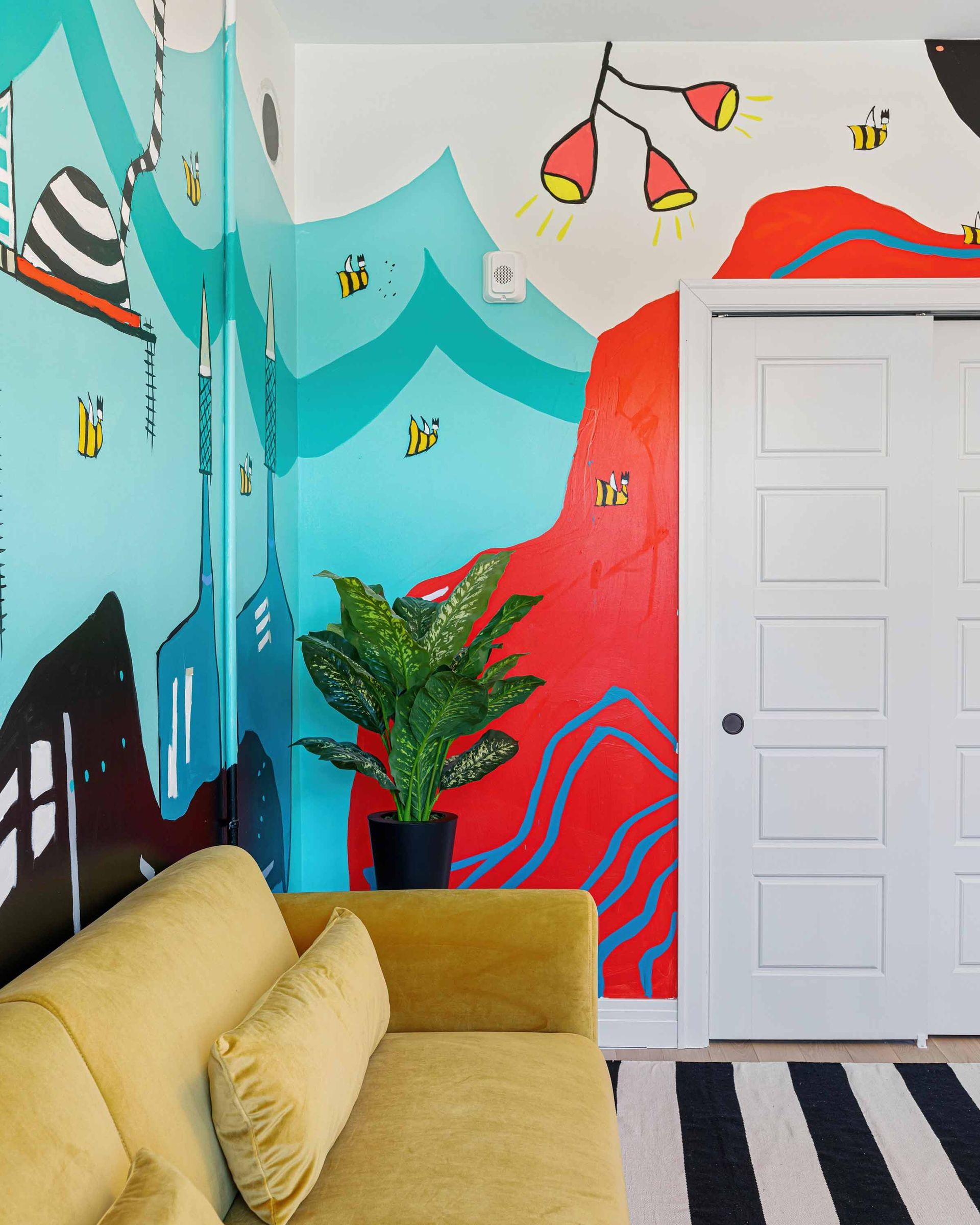Colorful mural-wall living room with yellow sofa, potted plant, striped rug, and white door