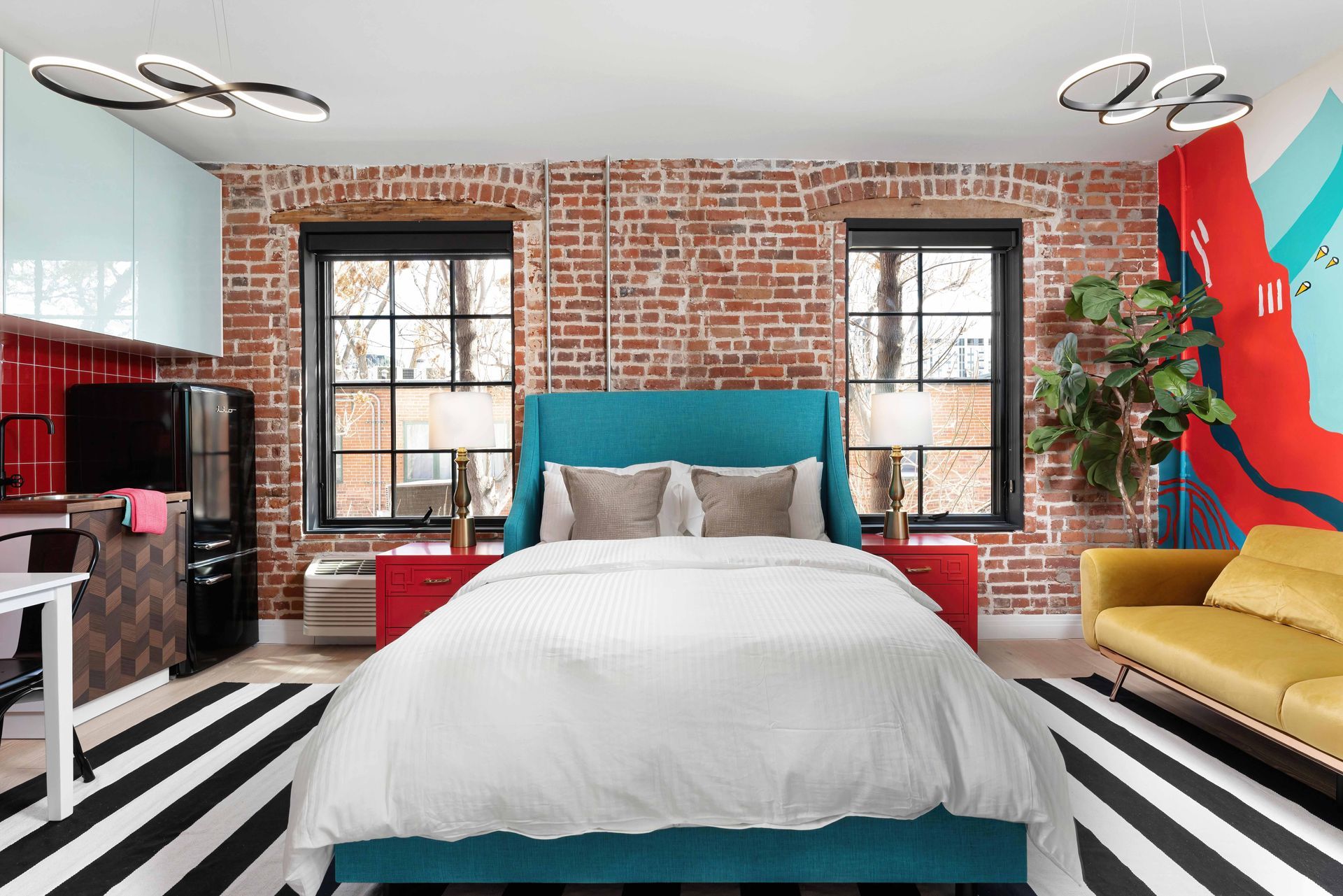 Modern bedroom with brick walls, teal bed, black-and-white rug, yellow sofa, and large windows