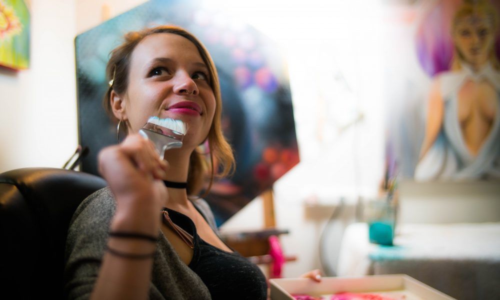 Woman smiling while painting in a colorful art studio, holding a brush near her face.