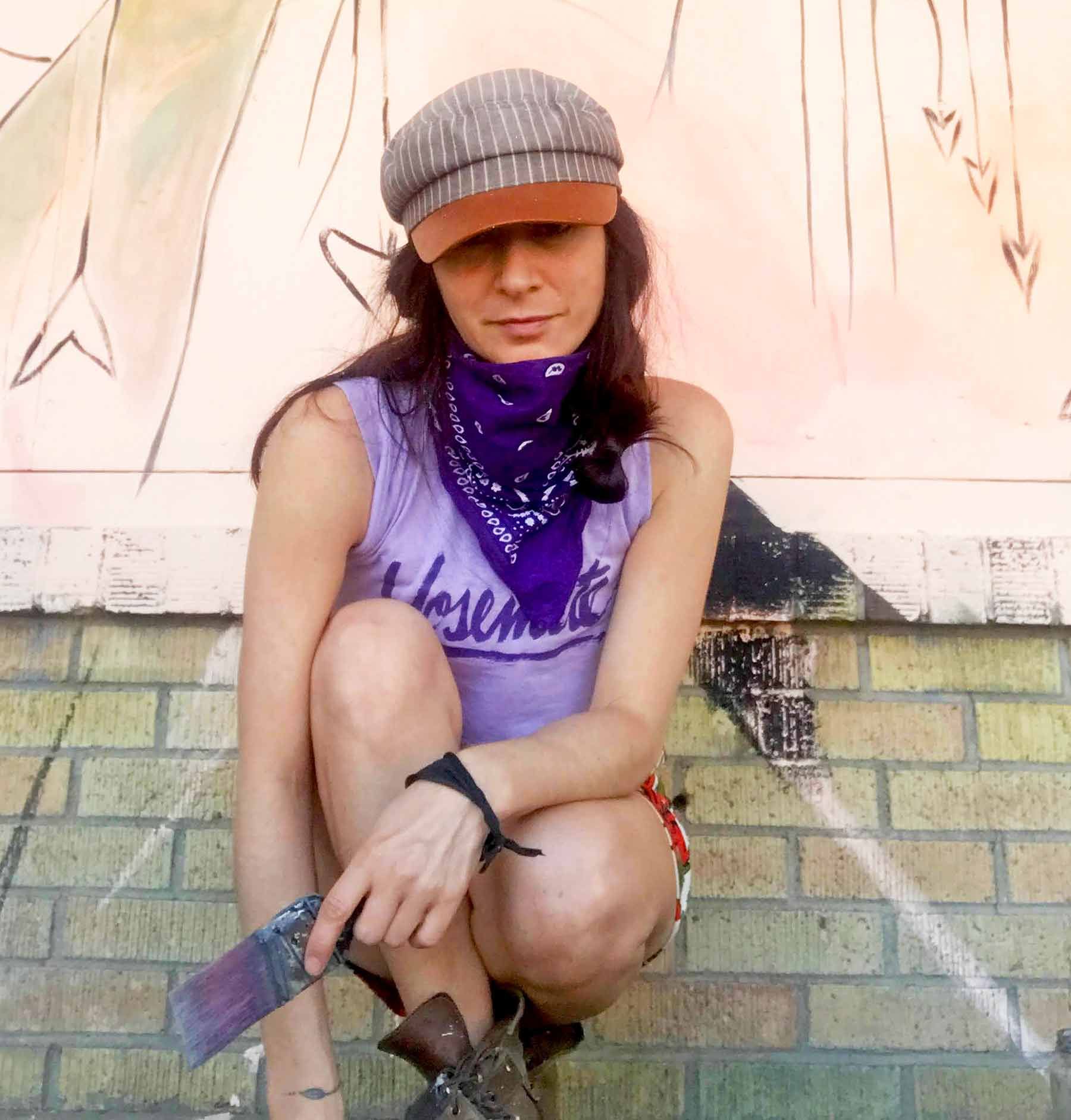 Person crouching outdoors against a mural, wearing a gray cap, purple bandana, and sleeveless shirt.