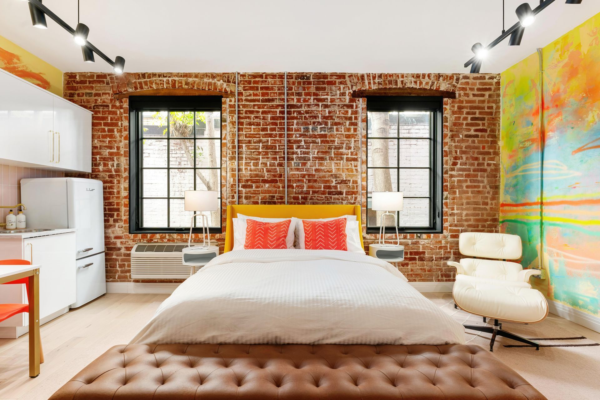 Modern loft bedroom with exposed brick walls, white bed, tufted bench, and colorful wall art