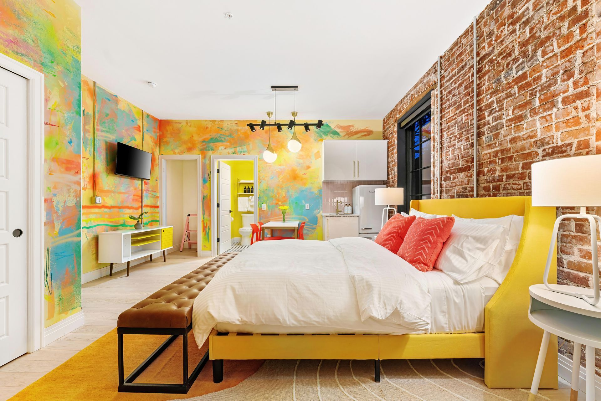 Colorful modern bedroom with brick wall, yellow bed, white bedding, and a wall-mounted TV.