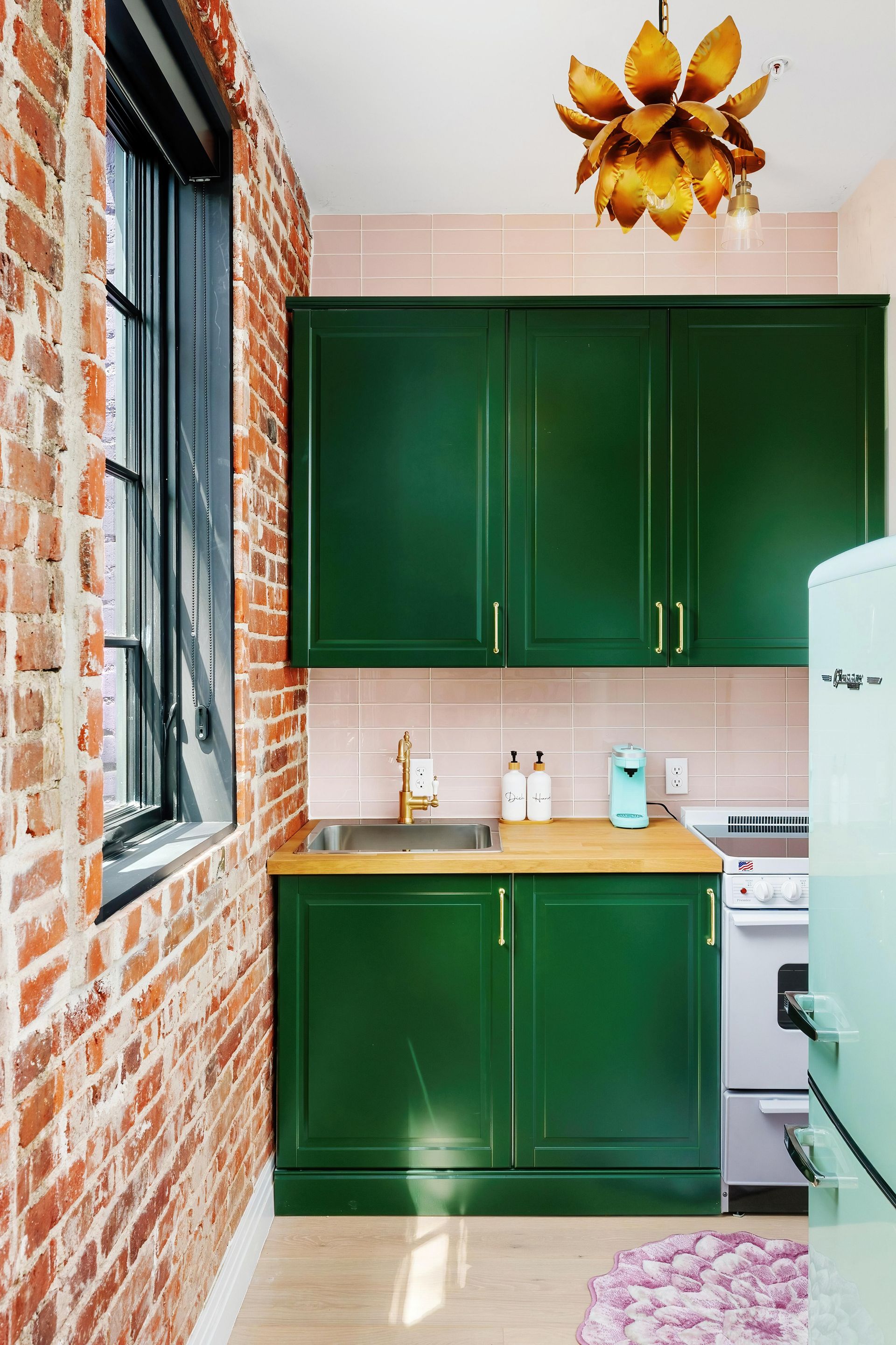 Small kitchen with green cabinets, brick wall, white stove and fridge, and sunburst wall decor.