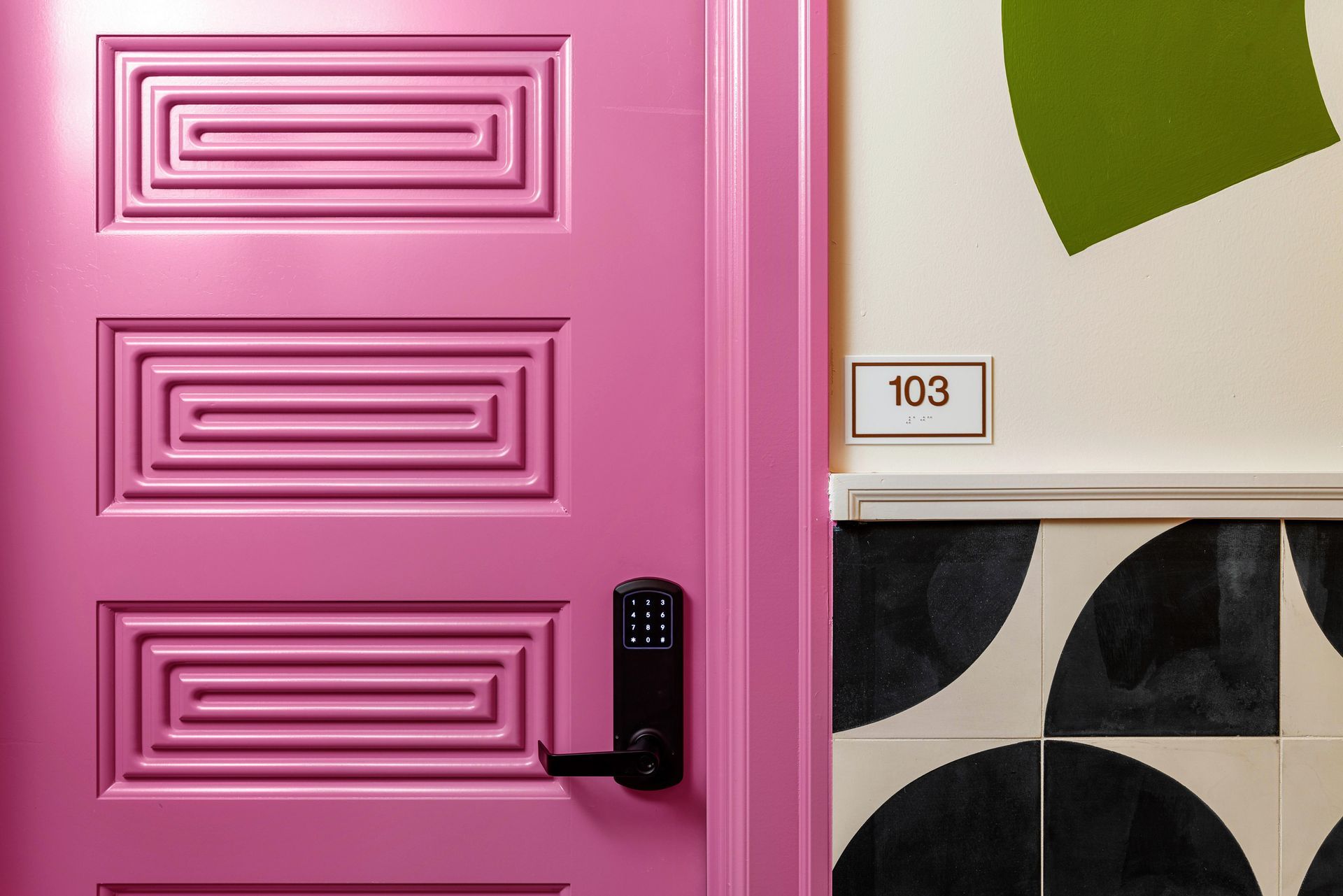 Pink front door with black handle beside tiled wall and small house number 103