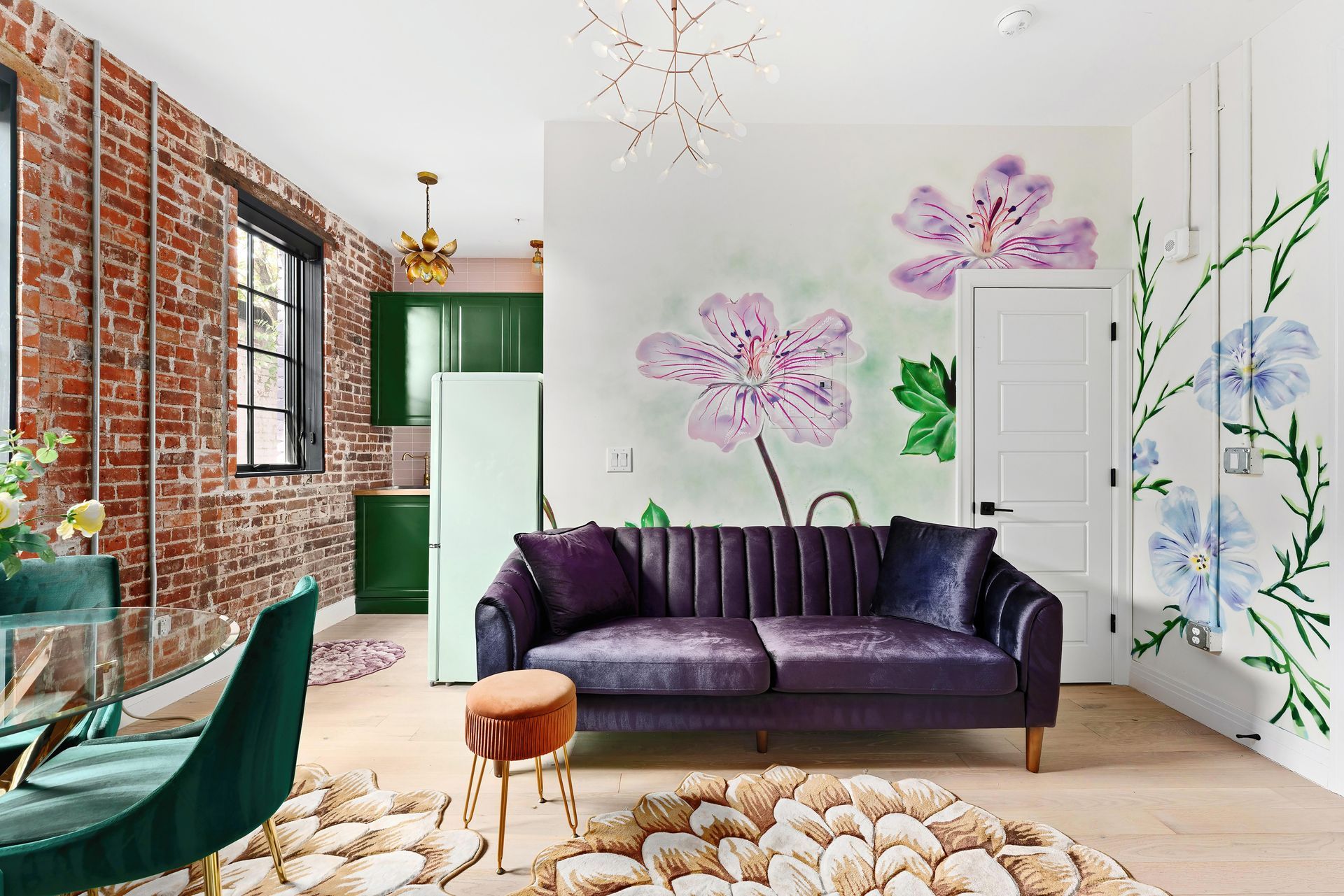 Bright living room with purple sofa, floral wall art, exposed brick, and green kitchen accents