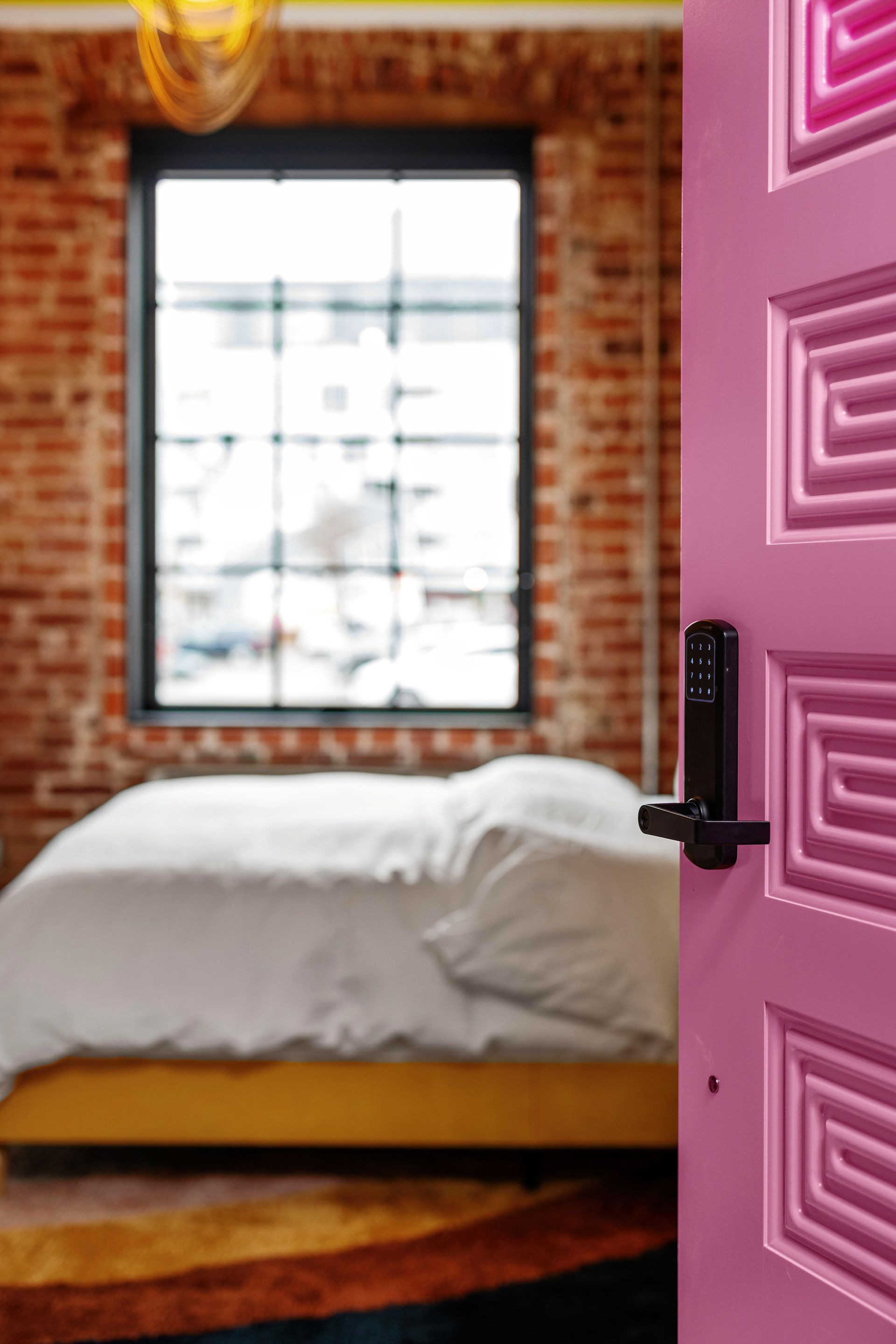 Bedroom seen through a partially open pink door with a black handle and a bed by a window.