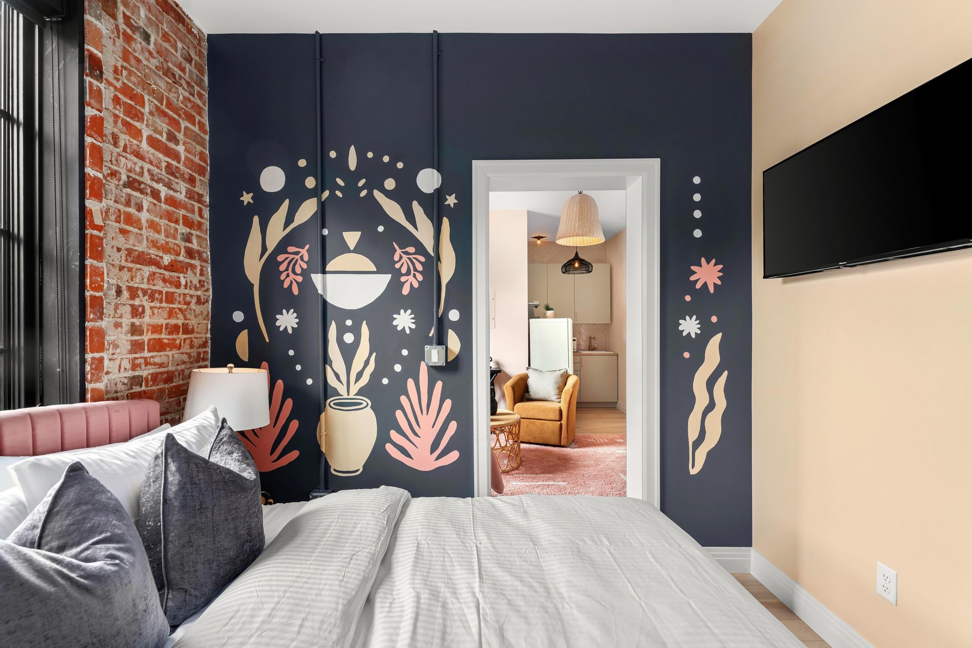 Bedroom with dark navy mural wall, brick accent, bed in foreground, and open doorway to a cozy sitting room
