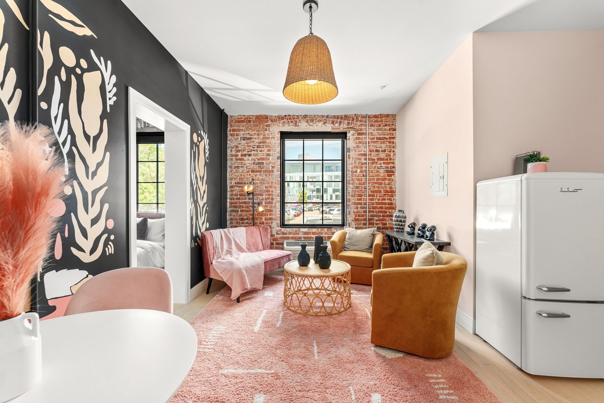 Bright living room with exposed brick wall, pink chair, mustard sofa, patterned rug, and white fridge