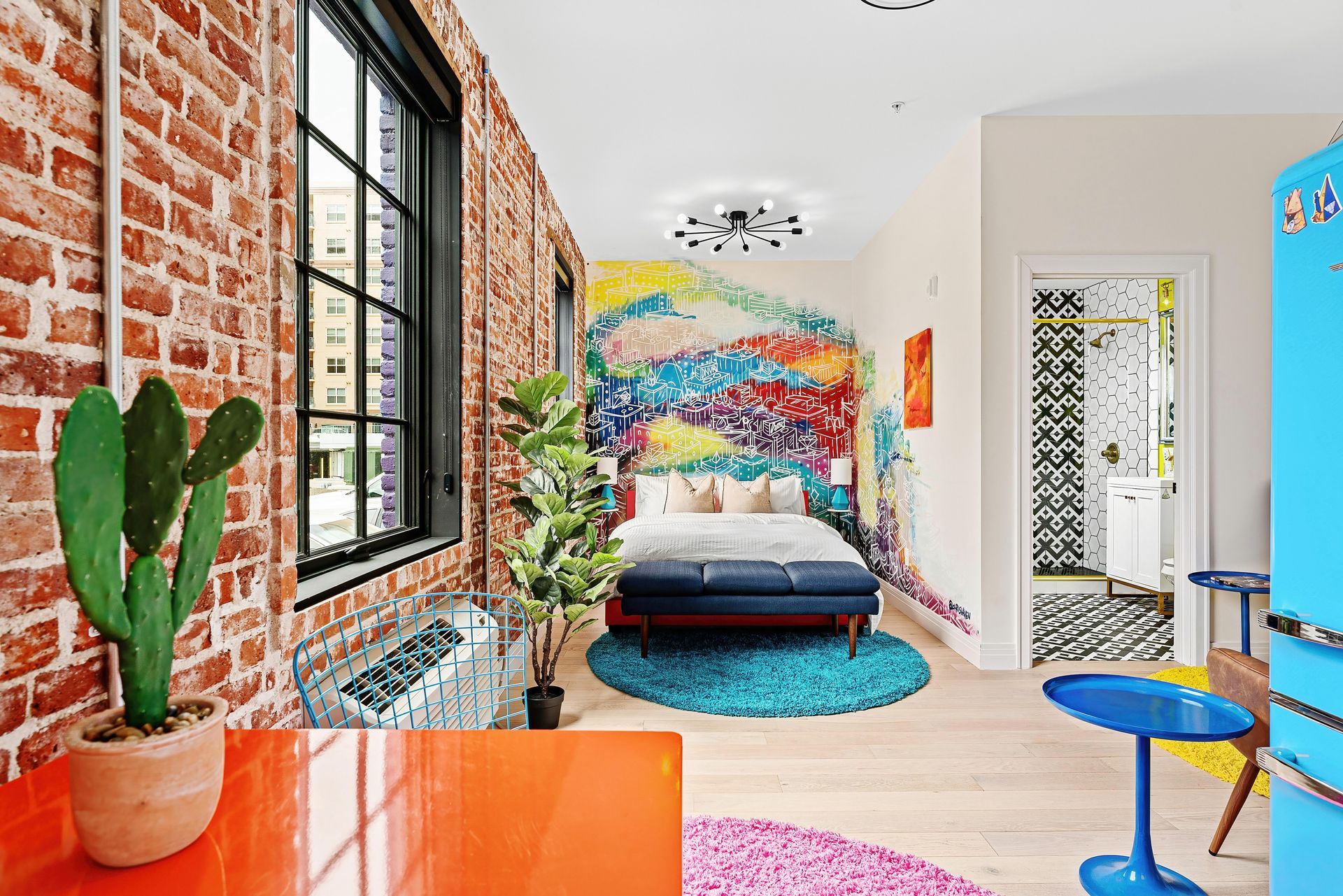 Bright loft bedroom with colorful mural, brick wall, bed, cactus, and orange desk by large windows