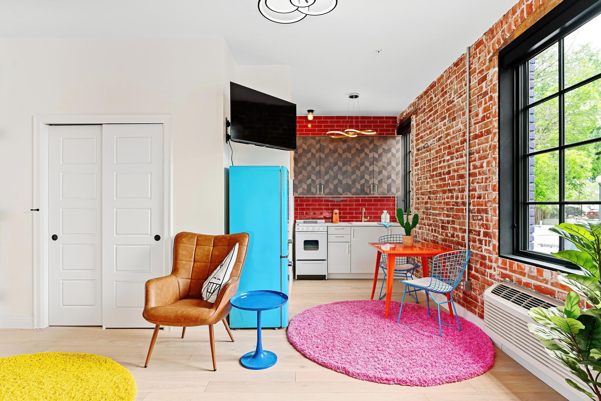 Bright modern room with brick wall, turquoise cabinet, pink rug, chair, TV, and large window