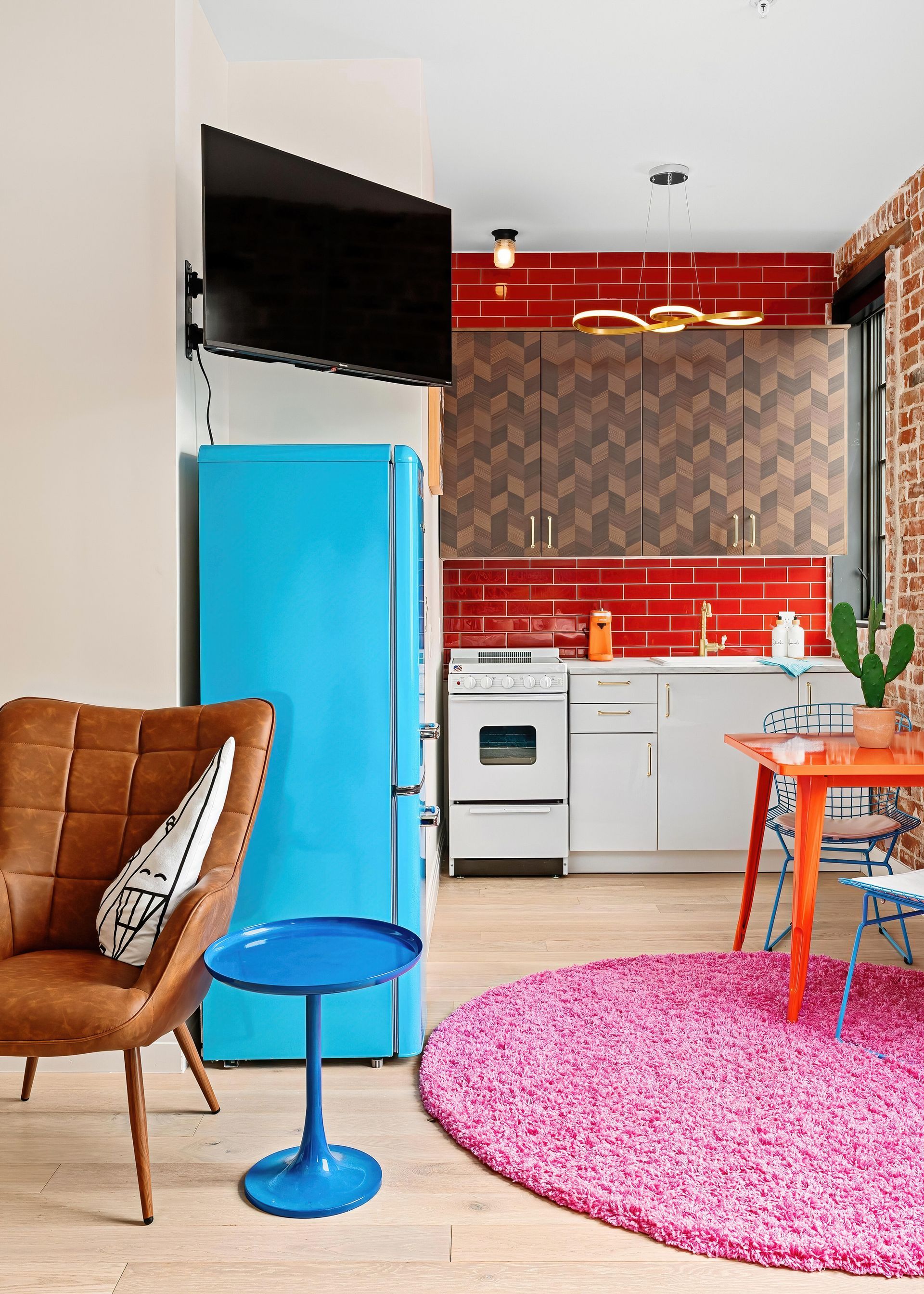 Bright modern kitchen-living room with red backsplash, blue fridge, white cabinets, and pink rug.
