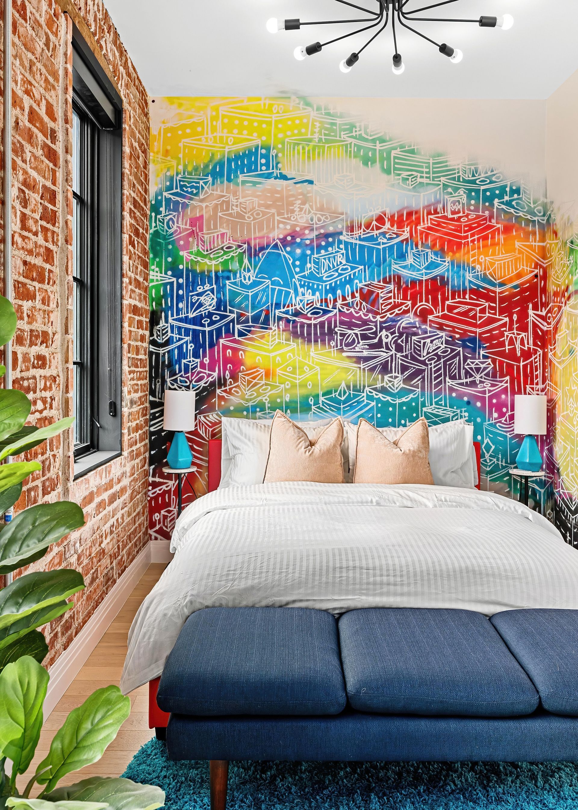 Colorful mural behind a neatly made bed in a bright modern bedroom with plants and a brick wall.