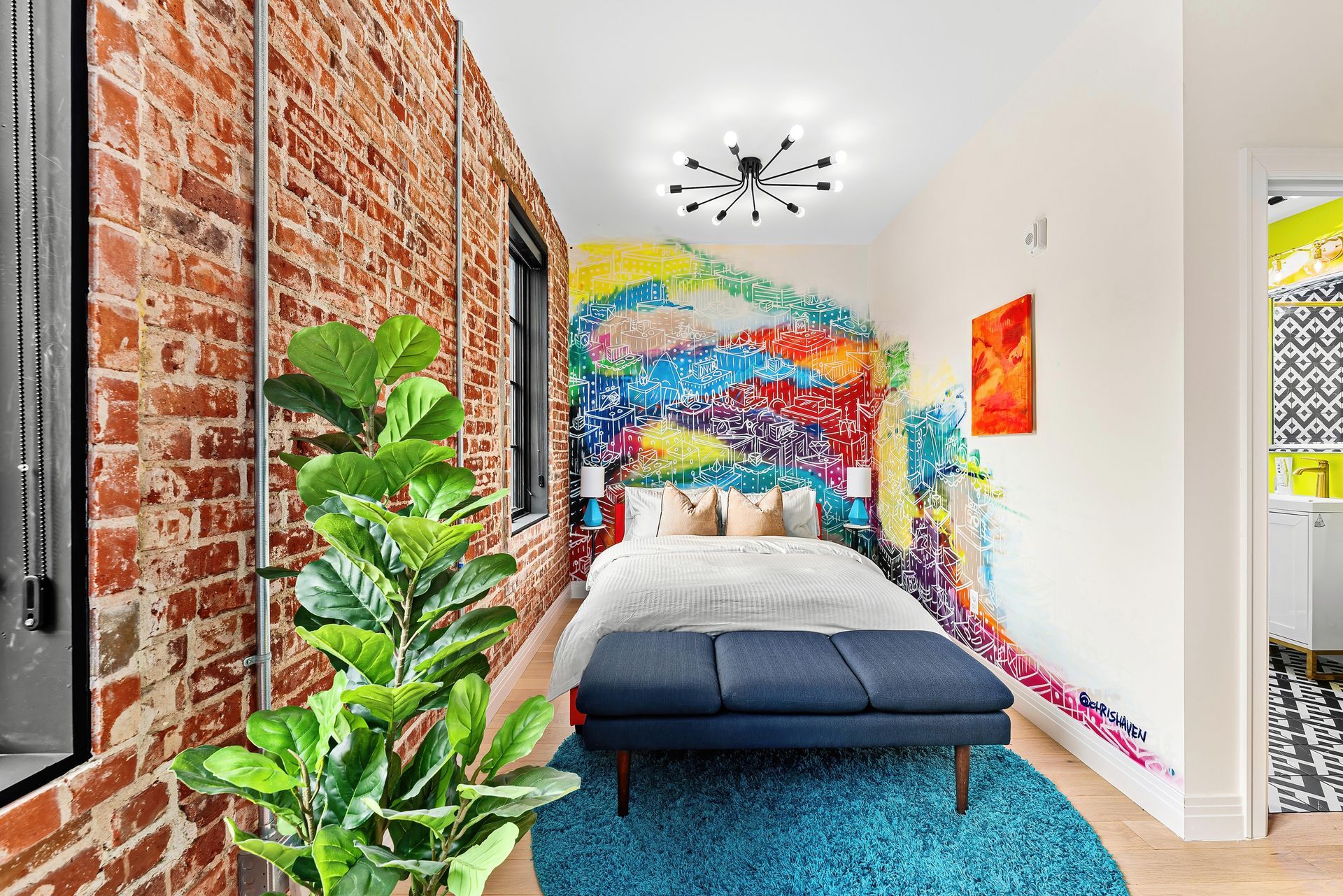 Narrow colorful bedroom with brick wall, teal rug, bed, large plant, and mural on the far wall