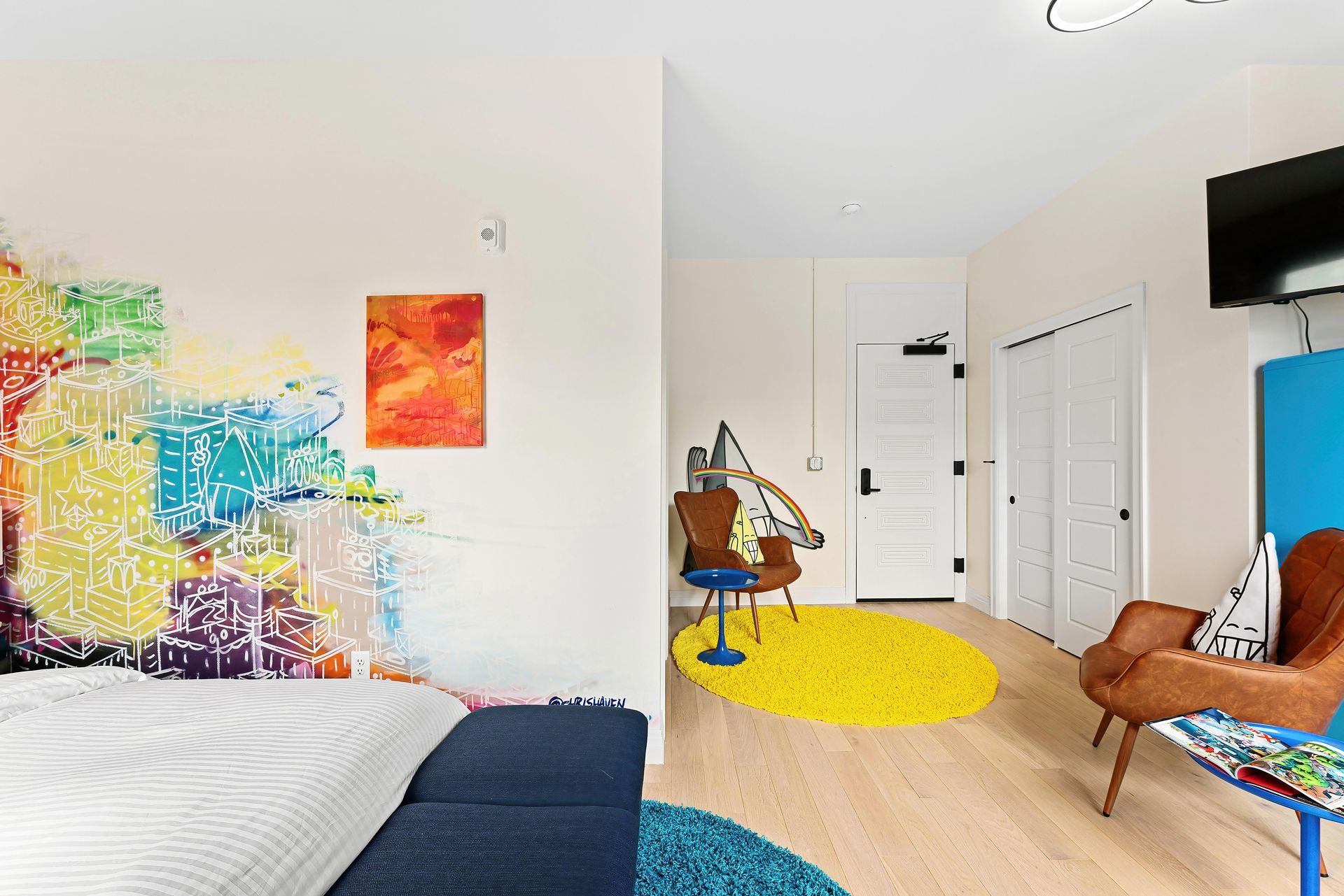 Bright bedroom with colorful wall art, white bed, blue chair, yellow rug, and a desk by the window.