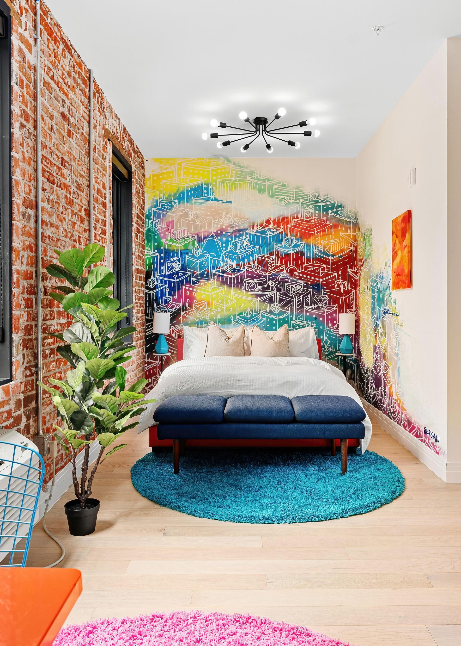 Modern bedroom with colorful graffiti-style accent wall, blue bed, turquoise rug, and plants