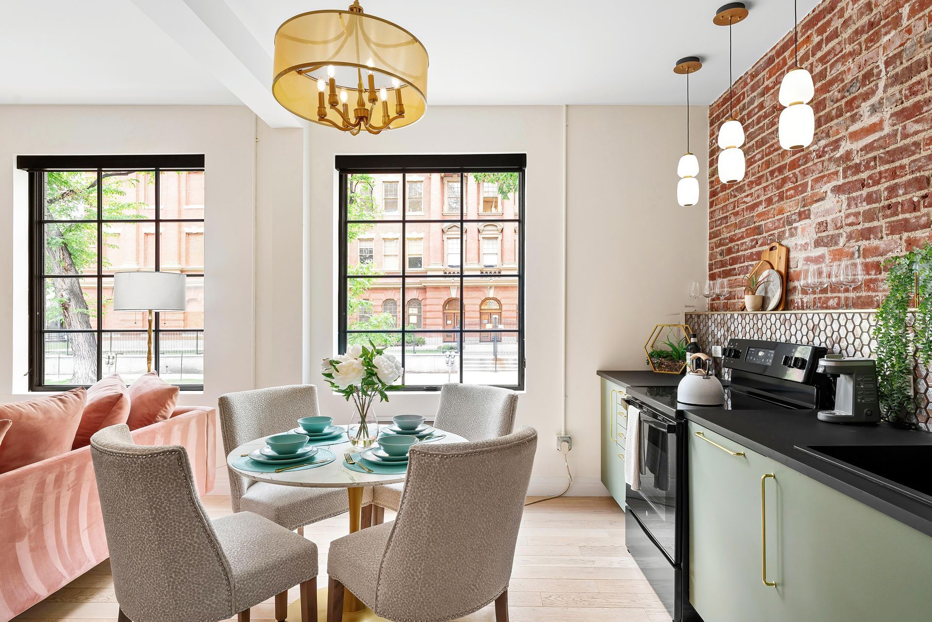 Bright living-dining room with pink sofa, round table, gold chandelier, brick wall, and black kitchen counter