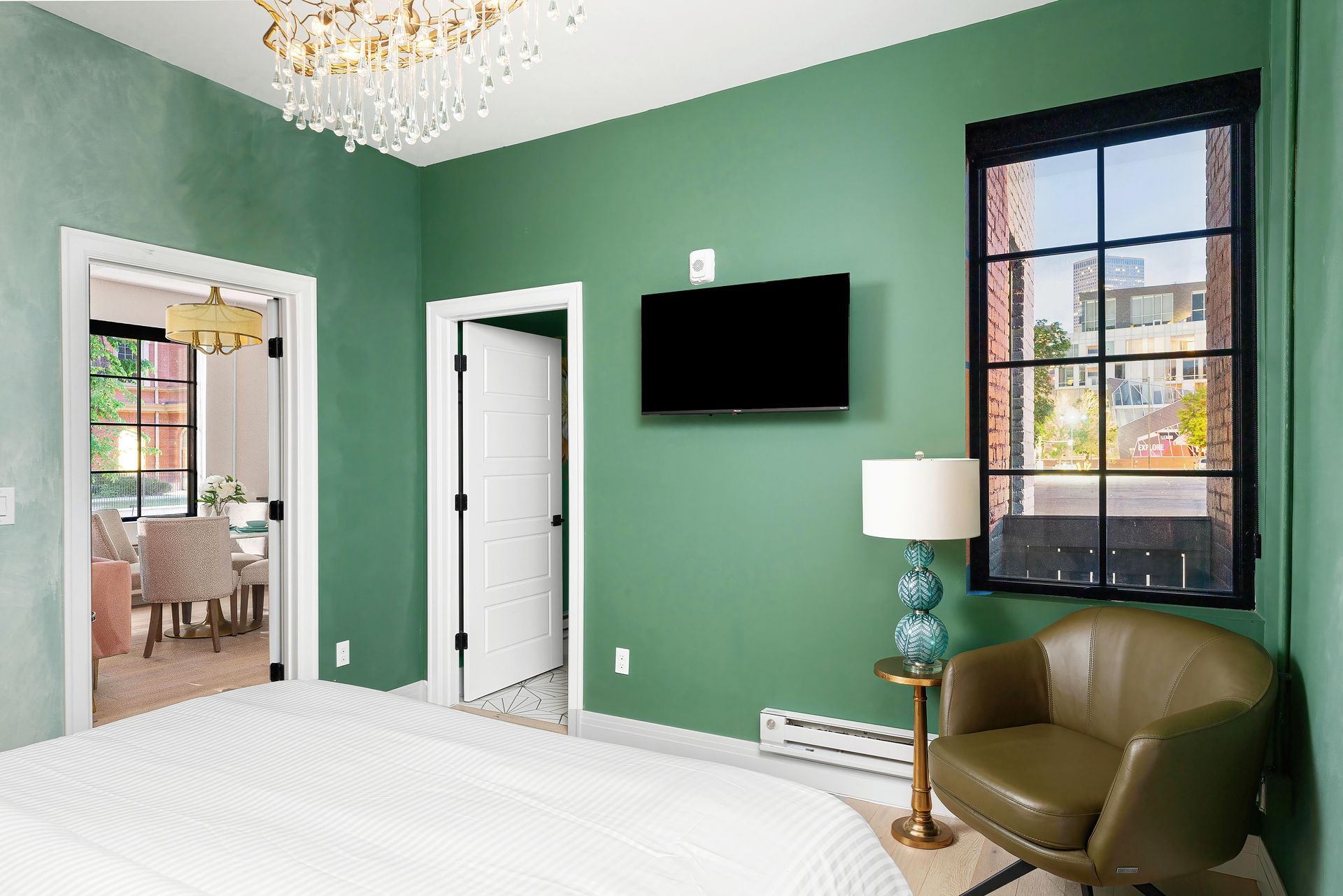 Green bedroom with wall-mounted TV, white door, chandelier, and olive chair near a window.