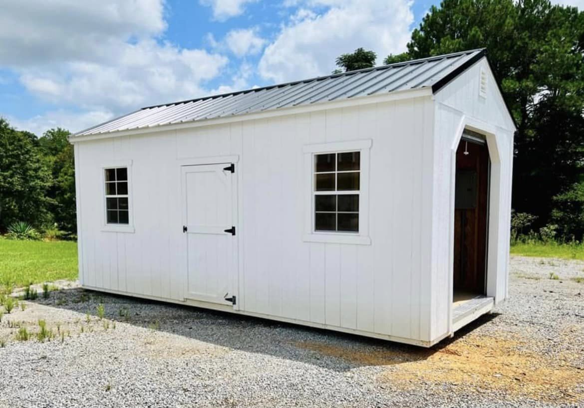 White shed with metal roof and windows on a gravel lot, with an open doorway.