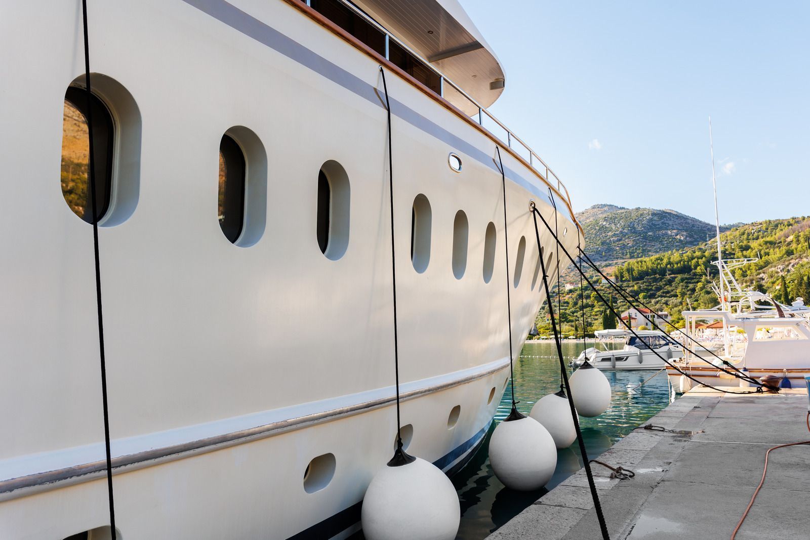 White yacht docked at a harbor, secured with black ropes and white buoys. Mountains in the background.