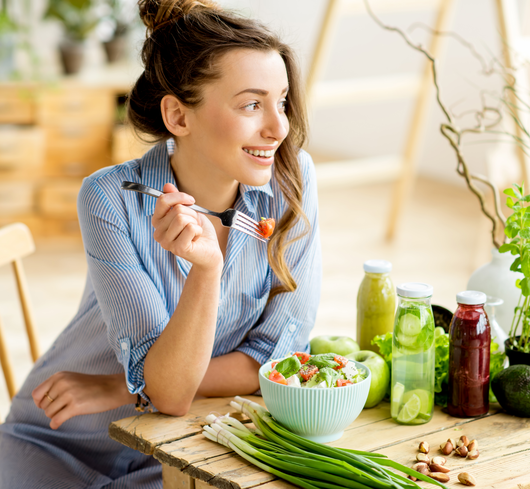 Woman smiling beside salad, green juices, and vegetables on a kitchen counter