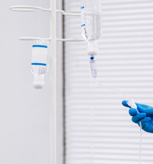 Gloved hand adjusting an IV drip line beside two hanging infusion bags in a bright clinic room