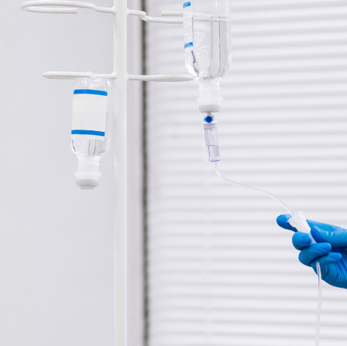 Gloved hand adjusting a clear IV drip line beside hanging infusion bags in a bright clinical room