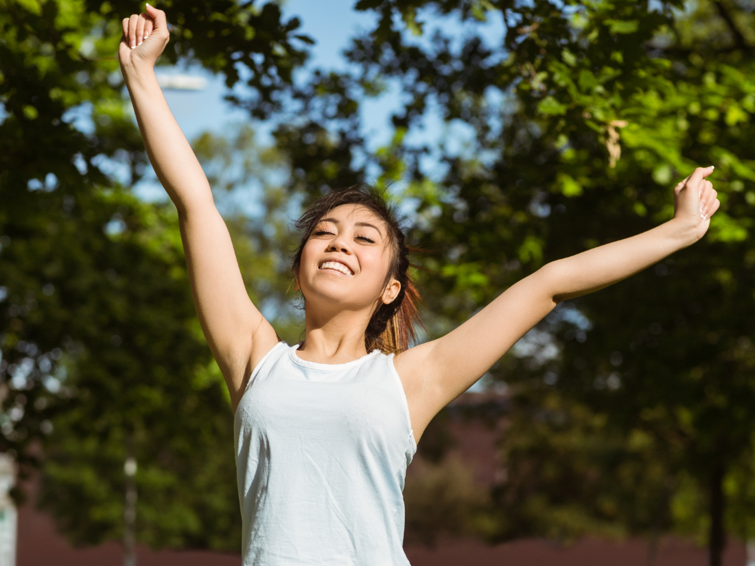Smiling woman with arms raised outdoors among trees on a sunny day