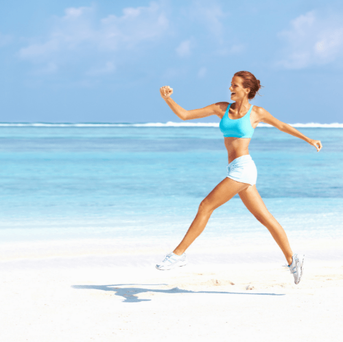 Woman jogging on a bright beach by the ocean, wearing a blue sports top and white shorts.