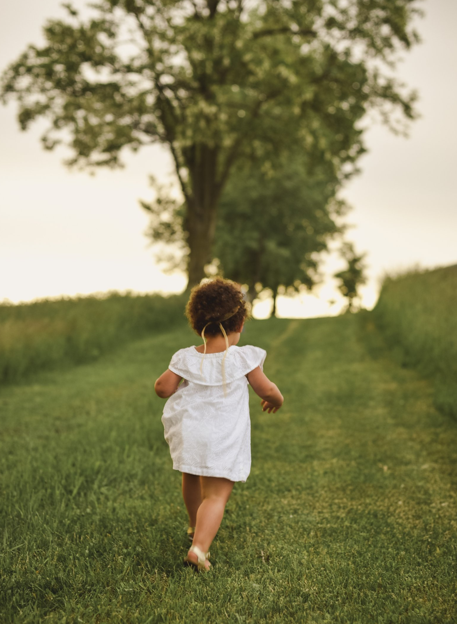 Child in a white dress running down a grassy path toward trees at sunset