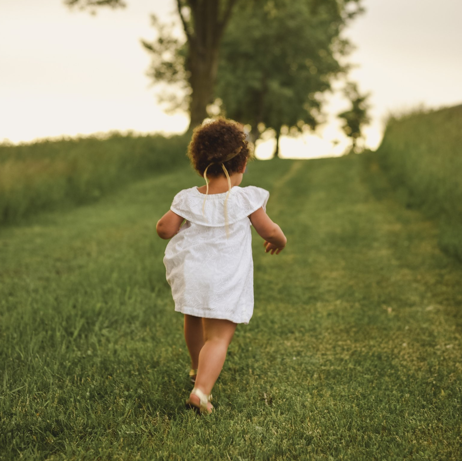 Child in a white dress running barefoot along a grassy path in a sunlit field