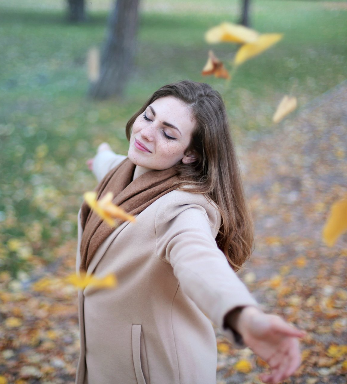 Woman in a beige coat smiling with outstretched arms in an autumn park, leaves falling around her.