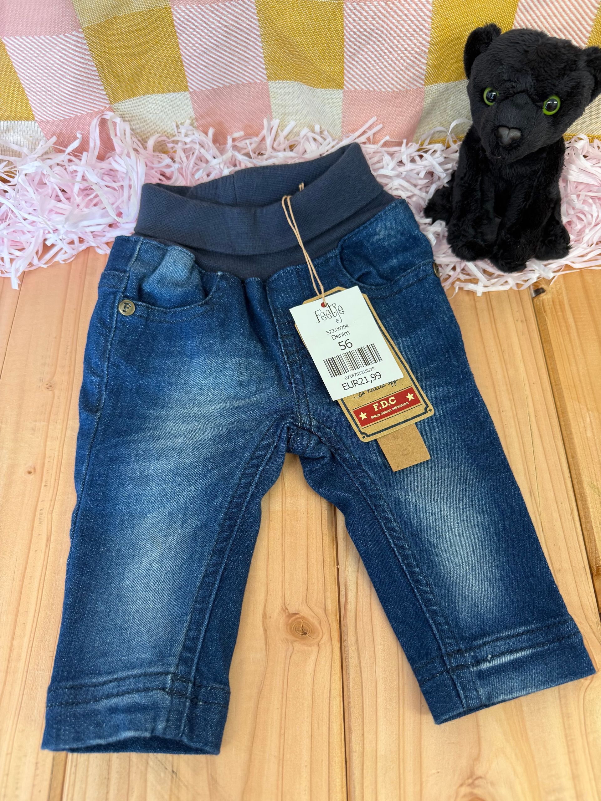 Een babyjeans met een label eraan ligt op een houten tafel naast een knuffeldier.