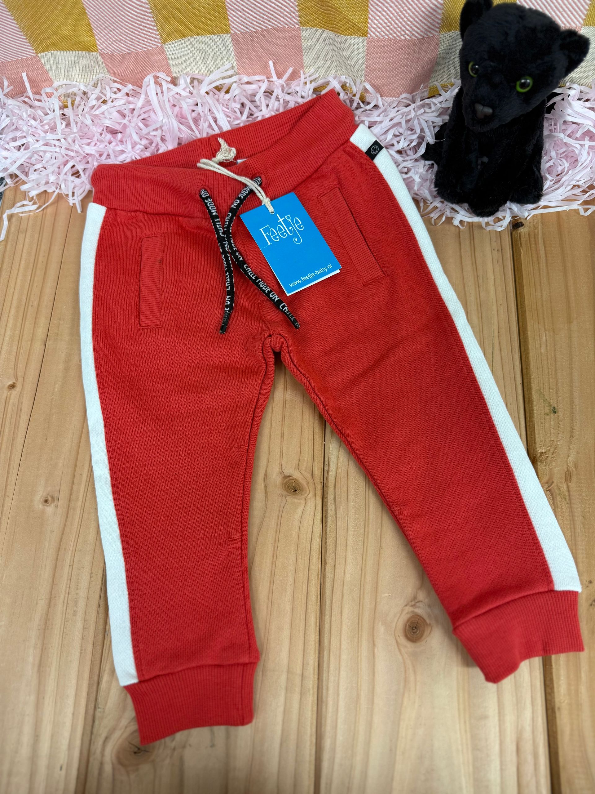 Een rode broek met witte strepen aan de zijkant ligt op een houten tafel naast een knuffeldier.