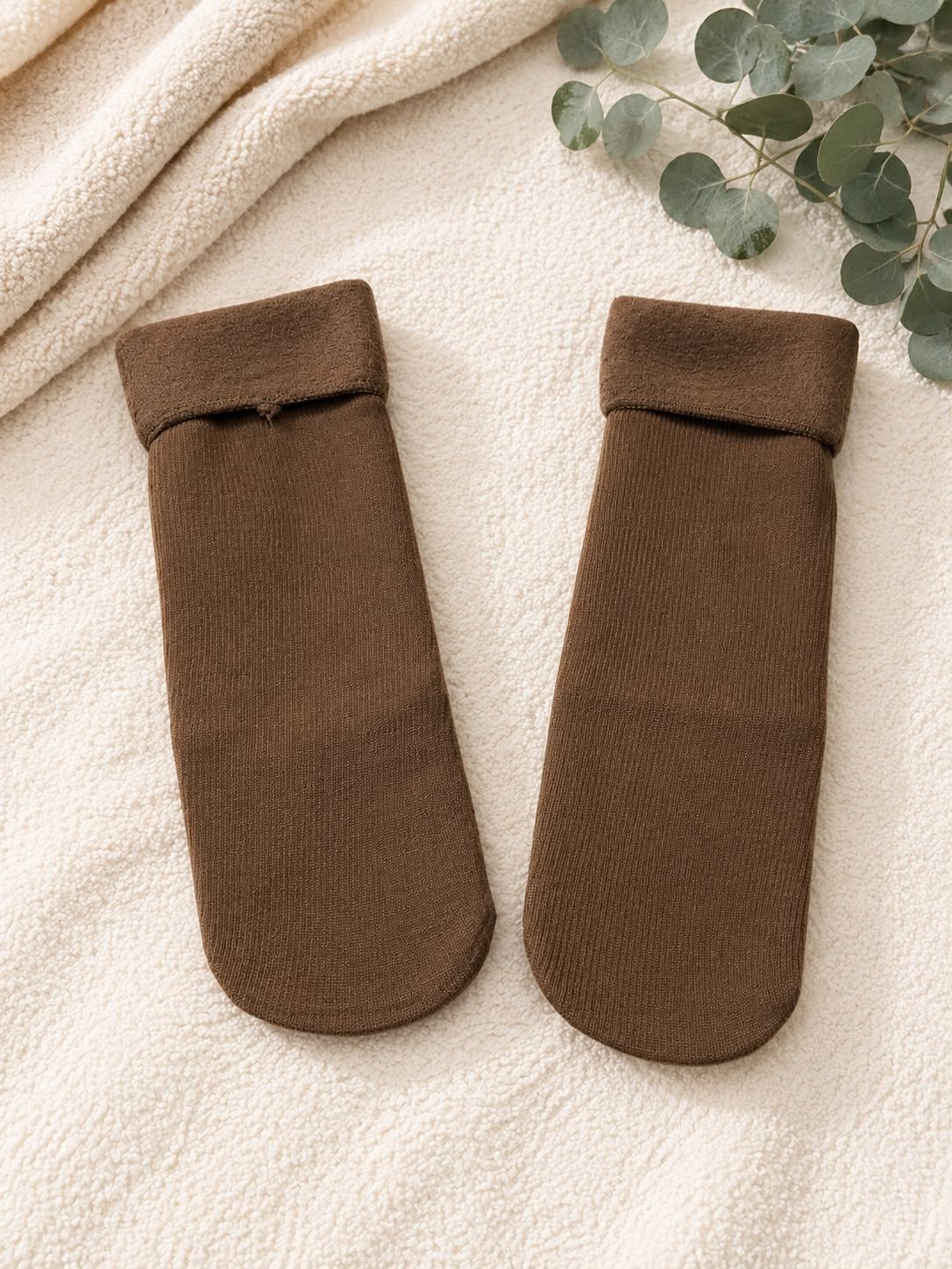 Un par de calcetines de punto marrones sobre un fondo de tela color crema con hojas de eucalipto cerca.