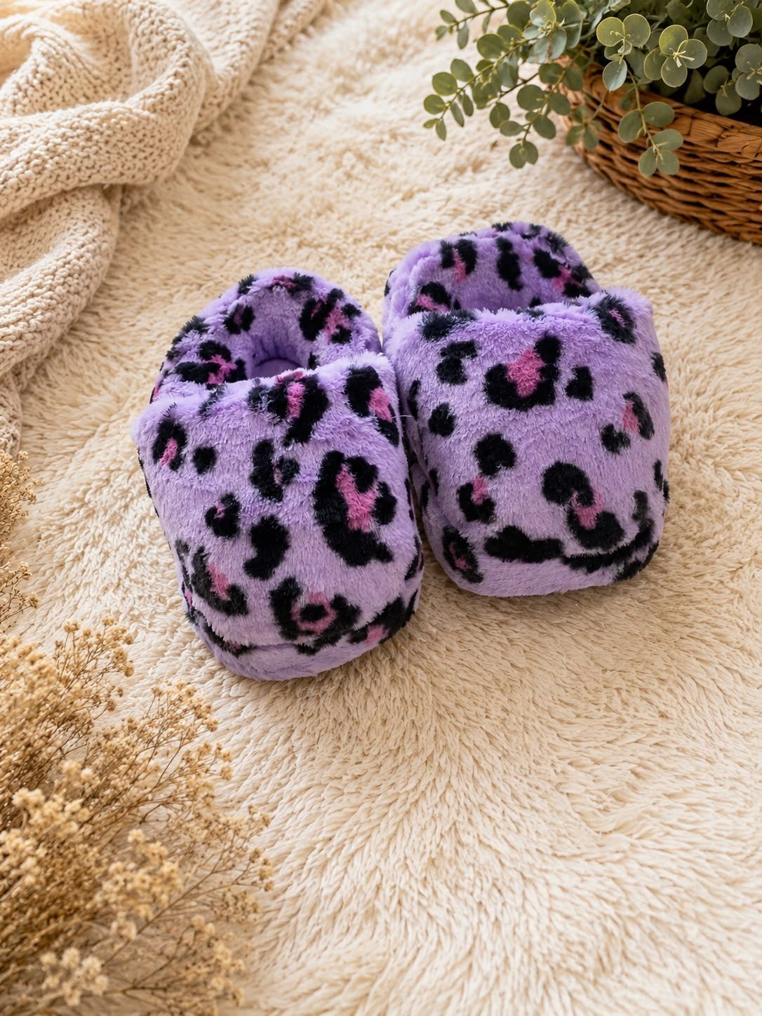 Pantuflas peludas con estampado de leopardo morado sobre una manta texturizada beige con una cesta de mimbre cerca.