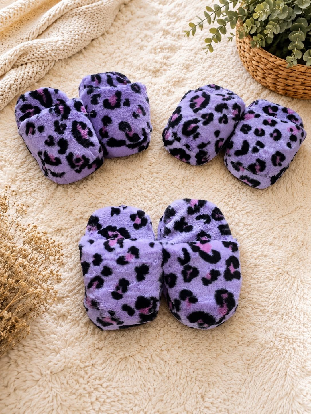 Pantuflas peludas con estampado de leopardo morado sobre una manta color crema junto a una cesta de mimbre y vegetación.