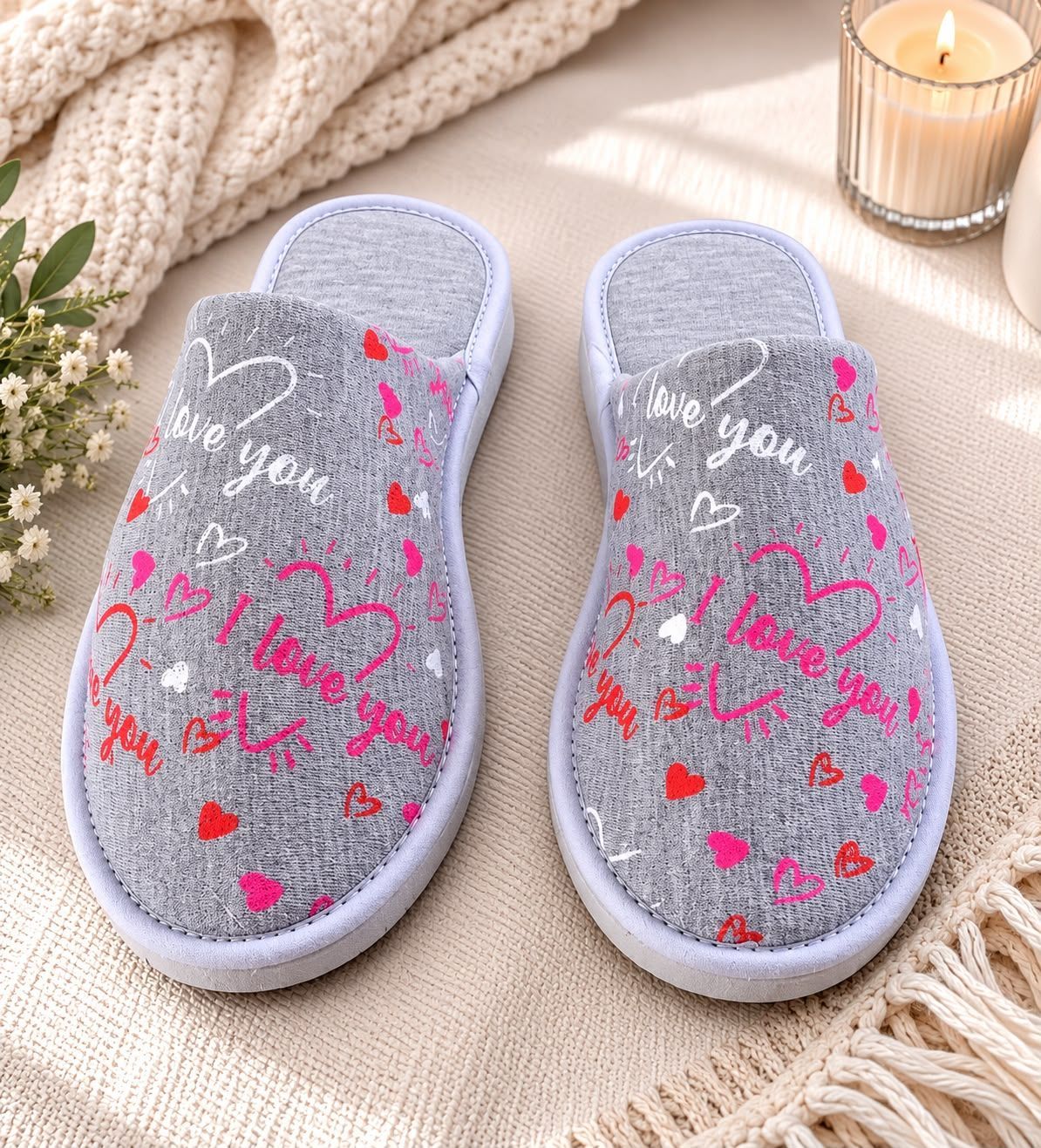 Pantuflas de punto grises con estampados de corazones rosas sobre una manta beige junto a una vela y una manta de punto.