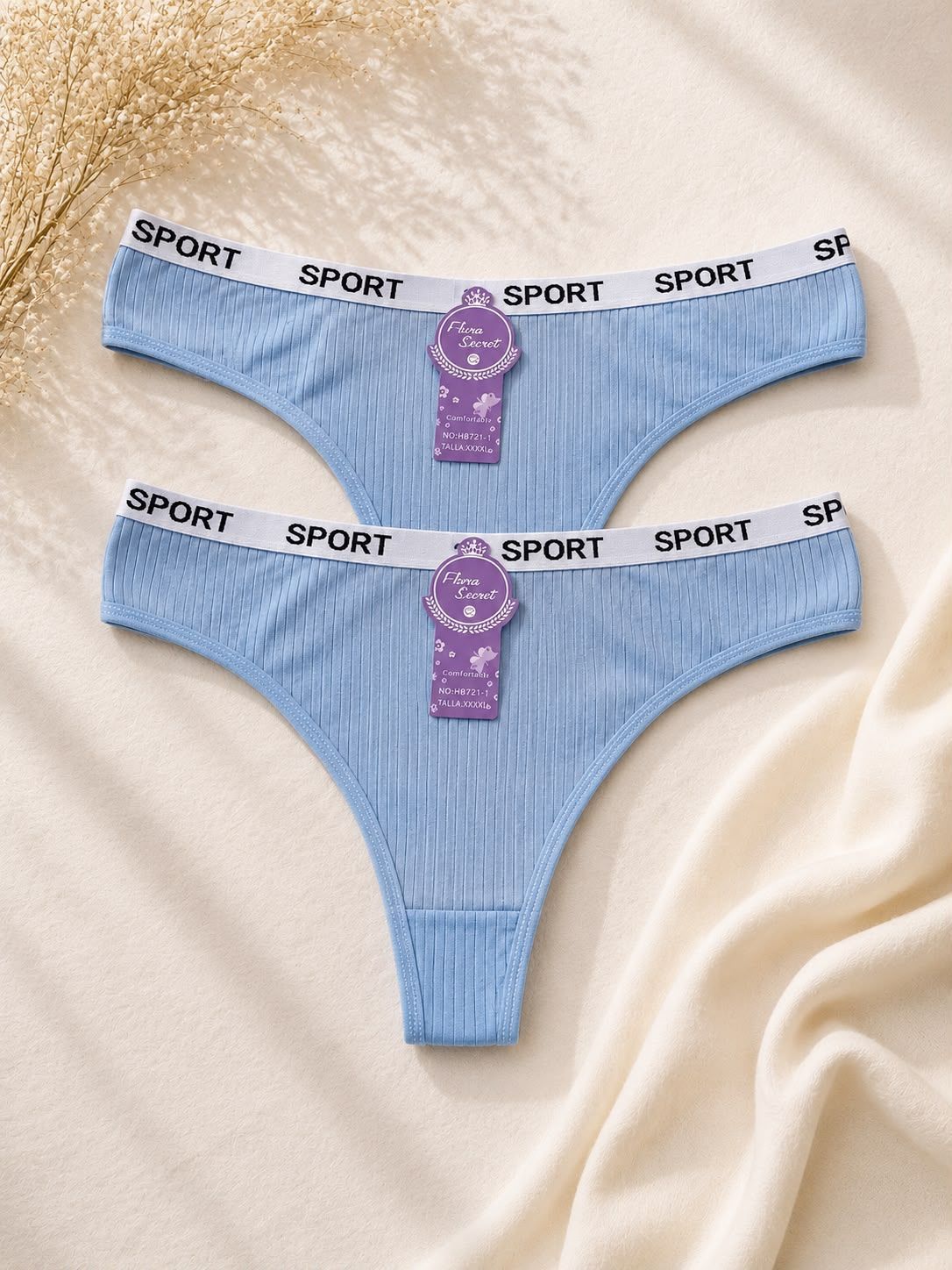 Dos tangas negras acanaladas con cinturillas blancas y moradas con la inscripción SPORT sobre un fondo de tela beige.
