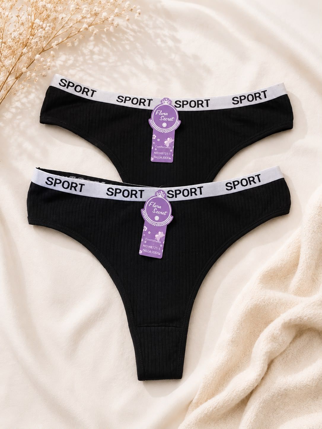 Dos tangas negras acanaladas con cinturillas blancas y moradas con la inscripción SPORT sobre un fondo de tela beige.