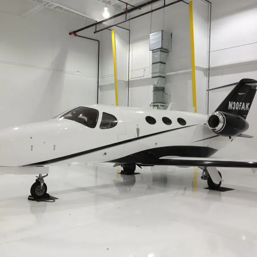 Small white private jet parked in a bright hangar, with black tail and yellow support straps.