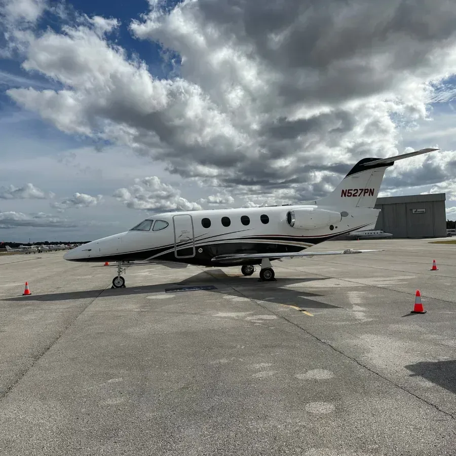 Small white private jet on a tarmac under cloudy sky, with orange cones nearby.
