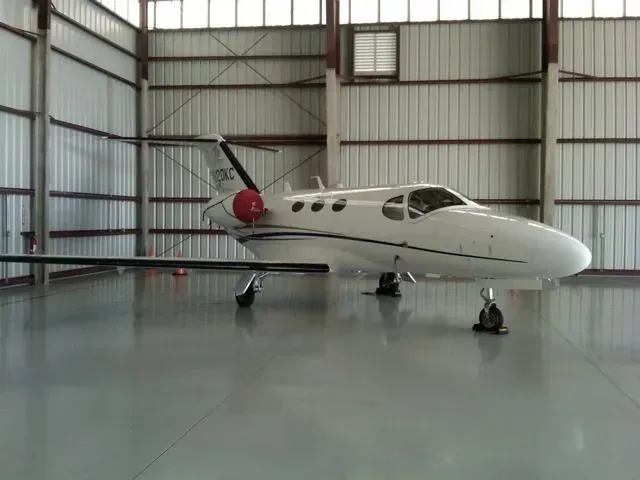Small white jet airplane parked inside a hangar with a red engine cover and hangar doors visible