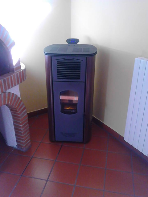 Stufa a pellet nera con una piccola fiamma in un angolo piastrellato tra un basamento in mattoni e un termosifone.